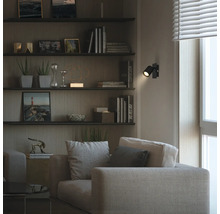 Salon moderne avec canapé en tissu et étagères murales. Un spot mural LED noir orientable crée un point lumineux dans la pièce sombre.