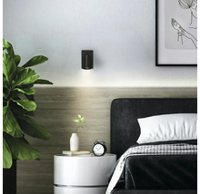 Applique murale cylindrique noire au-dessus d'un lit dans une chambre moderne et lumineuse avec table de chevet et plante.