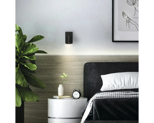 Applique murale cylindrique noire au-dessus d'un lit dans une chambre moderne et lumineuse avec table de chevet et plante.