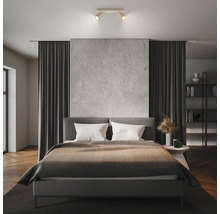 Chambre à coucher moderne avec lit rembourré gris, spot de plafond à deux lampes, panneau mural aspect béton, parquet et rideaux sombres.
