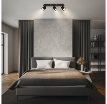 Chambre à coucher moderne avec un plafonnier à trois spots éclairant un lit gris, un mur aspect béton et des rideaux sombres.