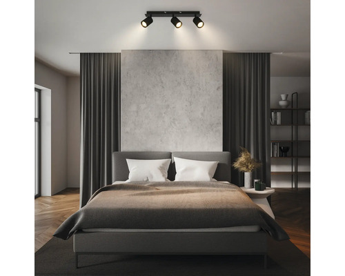 Chambre à coucher moderne avec un plafonnier à trois spots éclairant un lit gris, un mur aspect béton et des rideaux sombres.