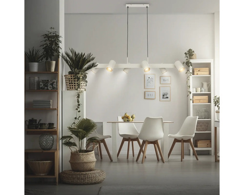 Salle à manger moderne et lumineuse avec une suspension à six lampes au-dessus d'une table avec quatre chaises, des étagères et des plantes d'intérieur.