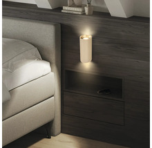 Applique murale cylindrique nervurée moderne sur un mur en bois foncé à côté d'un lit, diffusant de la lumière vers le haut et le bas.