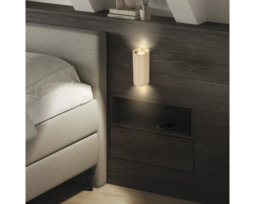 Applique murale cylindrique nervurée moderne sur un mur en bois foncé à côté d'un lit, diffusant de la lumière vers le haut et le bas.