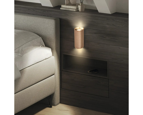 Applique murale cylindrique moderne avec une texture à lamelles verticales sur un panneau en bois sombre à côté d'un lit capitonné.
