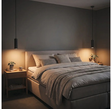 Chambre à coucher moderne avec lit capitonné, coussins gris, deux suspensions cylindriques noires et une table de chevet en bois.