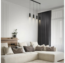 Salon moderne lumineux avec grand canapé en forme de L, coussins, rideaux sombres et suspension noire à quatre lampes cylindriques.