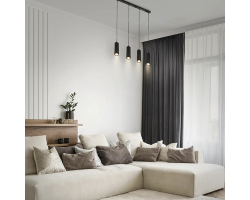 Salon moderne lumineux avec grand canapé en forme de L, coussins, rideaux sombres et suspension noire à quatre lampes cylindriques.