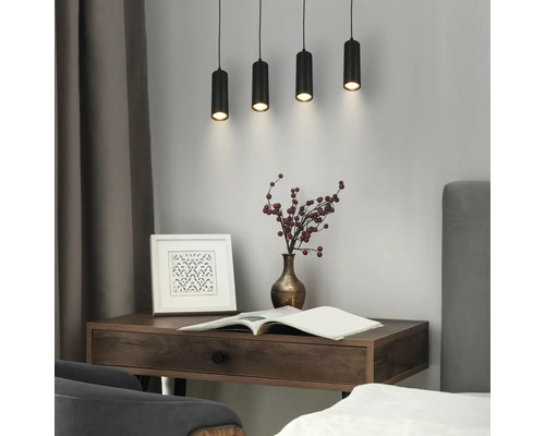 Quatre suspensions cylindriques noires au-dessus d'un bureau en bois dans une pièce lumineuse et organisée avec cadre, vase et livre.