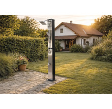 Moderne Solardusche aus Edelstahl im Garten mit Regenduschkopf, Handbrause und Fußbrause auf einem gepflasterten Weg neben einer Rasenfläche.