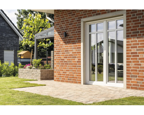 Porte-fenêtre blanche à croisillons dans un mur en briques rouges devant une terrasse pavée dans un jardin.