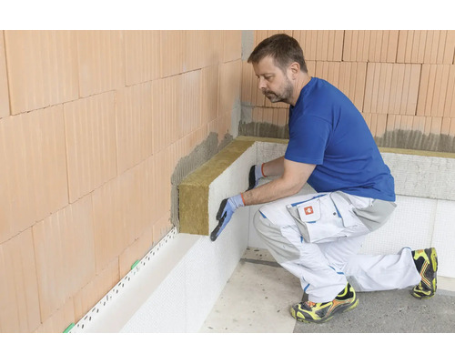 Un artisan pose des panneaux isolants en laine minérale sur un mur en briques pour une isolation thermique intérieure professionnelle.