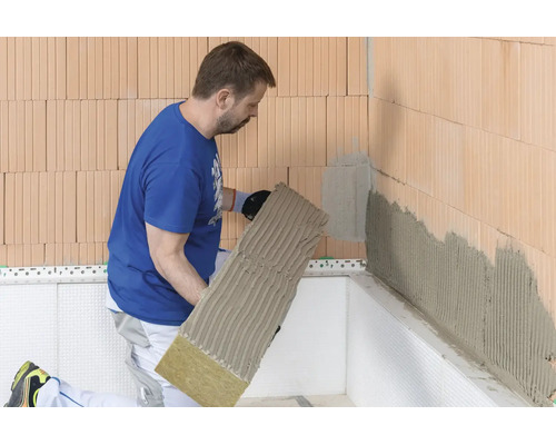 Artisan appliquant un panneau isolant en laine de roche avec du mortier-colle sur un mur en briques.