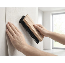 Une personne lisse du papier peint sur un mur avec une brosse de tapissier Rautner munie d'un manche en bois et de poils foncés.