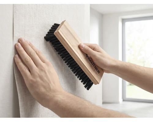 Une personne lisse du papier peint sur un mur avec une brosse de tapissier Rautner munie d'un manche en bois et de poils foncés.
