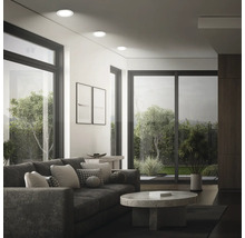 Salon moderne avec spots encastrés au plafond, grandes fenêtres, canapé et table basse ronde avec vue sur un jardin.