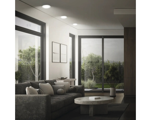 Salon moderne avec spots encastrés au plafond, grandes fenêtres, canapé et table basse ronde avec vue sur un jardin.