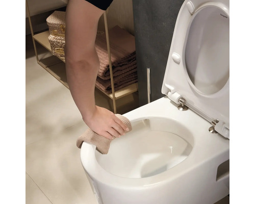 Une main nettoie le rebord de toilettes suspendues blanches avec un chiffon beige dans une salle de bains claire.
