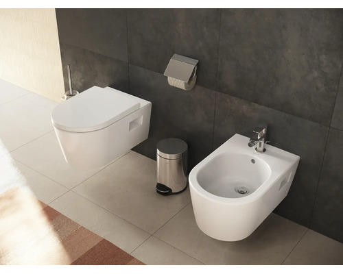 Salle de bains moderne avec WC suspendu, bidet avec robinetterie chromée, poubelle à pédale et porte-rouleau sur carrelage sombre.
