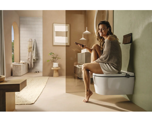 Femme assise sur des toilettes douche suspendues dans une salle de bains moderne et lumineuse, utilisant son smartphone.