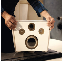 Mains tenant l'arrière d'une cuvette de WC suspendue en céramique blanche avec des ouvertures de montage et de raccordement.