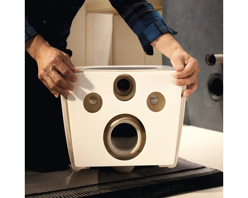Mains tenant l'arrière d'une cuvette de WC suspendue en céramique blanche avec des ouvertures de montage et de raccordement.