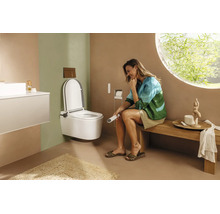 Salle de bains moderne avec WC lavant suspendu, femme assise sur un banc en bois tenant une télécommande et une fenêtre ronde.