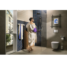 Salle de bain moderne avec personne en peignoir, douche à l'italienne, paroi en verre, WC suspendu, porte-serviettes et grande fenêtre.