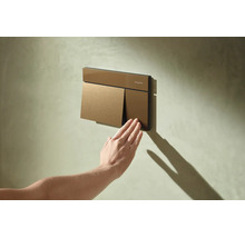 Plaque de déclenchement Hansgrohe pour toilettes sur un mur texturé, une main appuie sur le petit bouton de chasse d'eau.