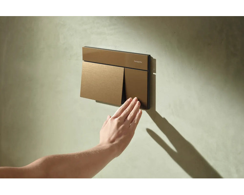 Plaque de déclenchement Hansgrohe pour toilettes sur un mur texturé, une main appuie sur le petit bouton de chasse d'eau.