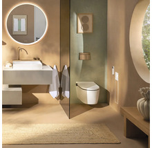 Salle de bains moderne avec vasque à poser blanche, miroir rond lumineux, WC suspendu et tapis naturel dans des tons terreux.