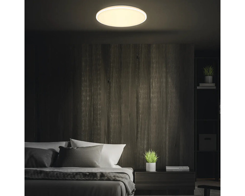 Runde LED-Deckenleuchte beleuchtet in einem dunklen Schlafzimmer mit holzgetäfelten Wänden, einem Bett und einem Nachttisch mit Pflanze.