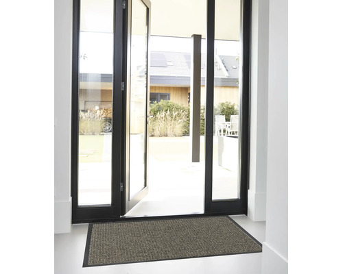 Paillasson structuré avec bordure noire dans une entrée lumineuse devant une porte vitrée ouverte avec un cadre noir.