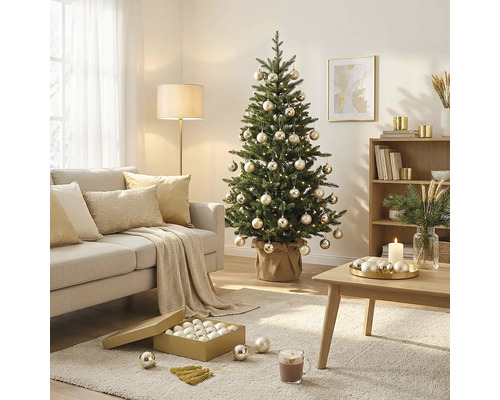 Sapin de Noël décoré de boules et d'une guirlande lumineuse dans un salon lumineux avec canapé, table basse et tapis.