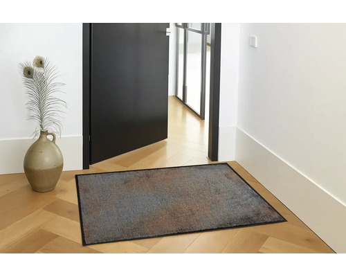 Paillasson rectangulaire multicolore avec un bord noir posé sur un parquet en bois clair dans une entrée lumineuse à côté d'un vase.