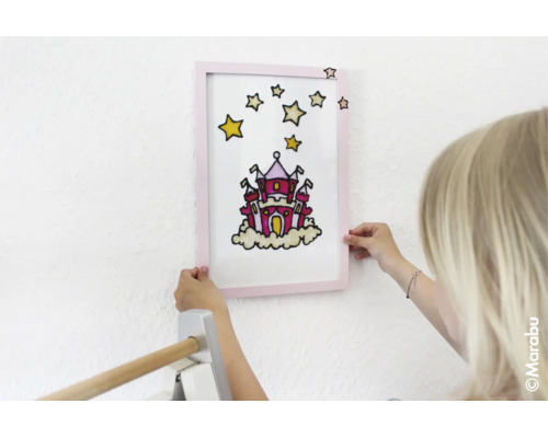 Kind hält ein gerahmtes Bild mit einem Schloss und Sternen an eine Wand.