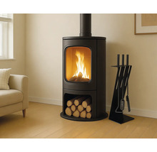 Poêle à bois noir moderne avec grande vitre, feu brûlant, compartiment à bûches et serviteur de cheminée dans un salon lumineux.
