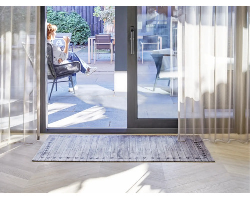 Tapis rectangulaire avec motif de planches de bois grises et bordure décorative dans un salon lumineux devant une baie vitrée coulissante.