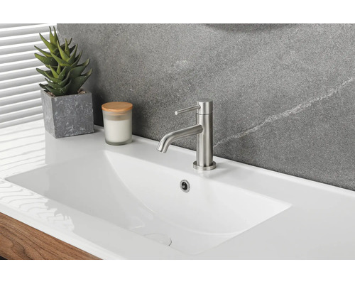Lavabo moderne en céramique blanche avec mitigeur monocommande en métal brossé, plante grasse et bougie devant un mur en pierre.