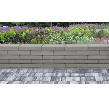 Mur de jardin en blocs de béton lisses et gris devant un parterre de fleurs et de graminées, adjacent à une surface pavée.