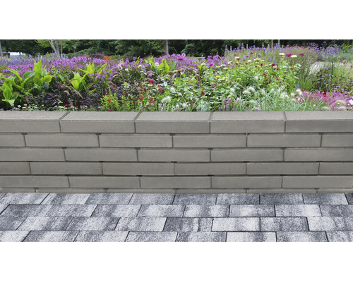 Mur de jardin en blocs de béton lisses et gris devant un parterre de fleurs et de graminées, adjacent à une surface pavée.