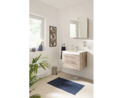 Salle de bain lumineuse avec meuble sous-lavabo suspendu aspect bois, lavabo blanc, armoire de toilette éclairée et tapis de bain bleu.