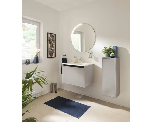 Salle de bains moderne avec meuble vasque blanc, miroir rond et meuble latéral. Inclut un tapis de bain et une poubelle.
