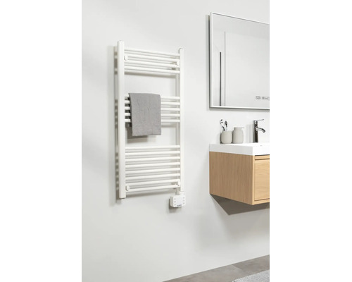 Salle de bains moderne avec radiateur sèche-serviettes électrique blanc, serviette grise, meuble sous-vasque en bois et grand miroir.