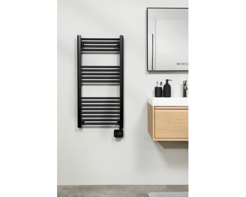 Sèche-serviettes électrique moderne noir avec thermostat numérique, monté à côté d'un meuble sous-vasque en bois dans une salle de bains claire.