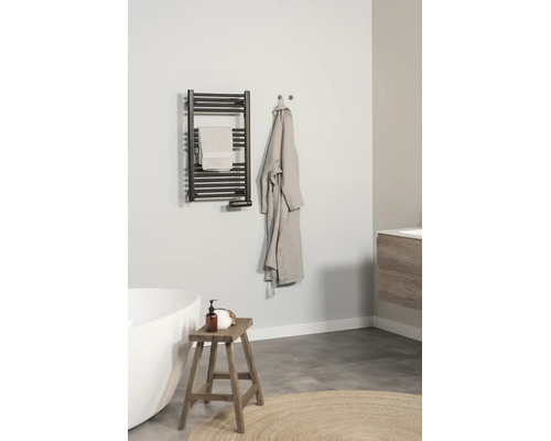 Salle de bains moderne et lumineuse avec un radiateur sèche-serviettes noir au mur, une baignoire blanche, un tabouret en bois et un peignoir.