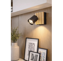 Spot mural moderne avec une tête cylindrique noire et une base en lattes de bois clair, monté sur un mur en bois au-dessus de cadres photo.