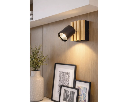 Spot mural moderne avec une tête cylindrique noire et une base en lattes de bois clair, monté sur un mur en bois au-dessus de cadres photo.