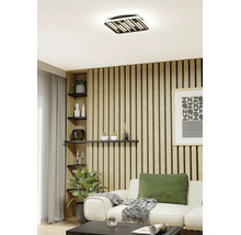 Moderne LED Deckenleuchte in quadratischer Form mit Elementen in Holzoptik in einem hellen Wohnzimmer mit Holzpaneelwand.